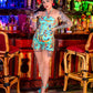 Woman in a colorful dress standing in a bar with neon lights and decor.
