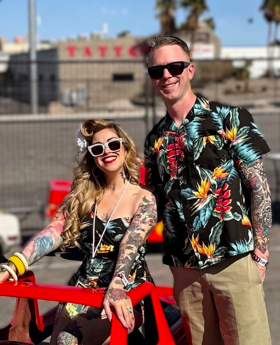Two people with tattoos posing outdoors, one in a floral shirt and sunglasses, the other in a black dress with floral patterns.