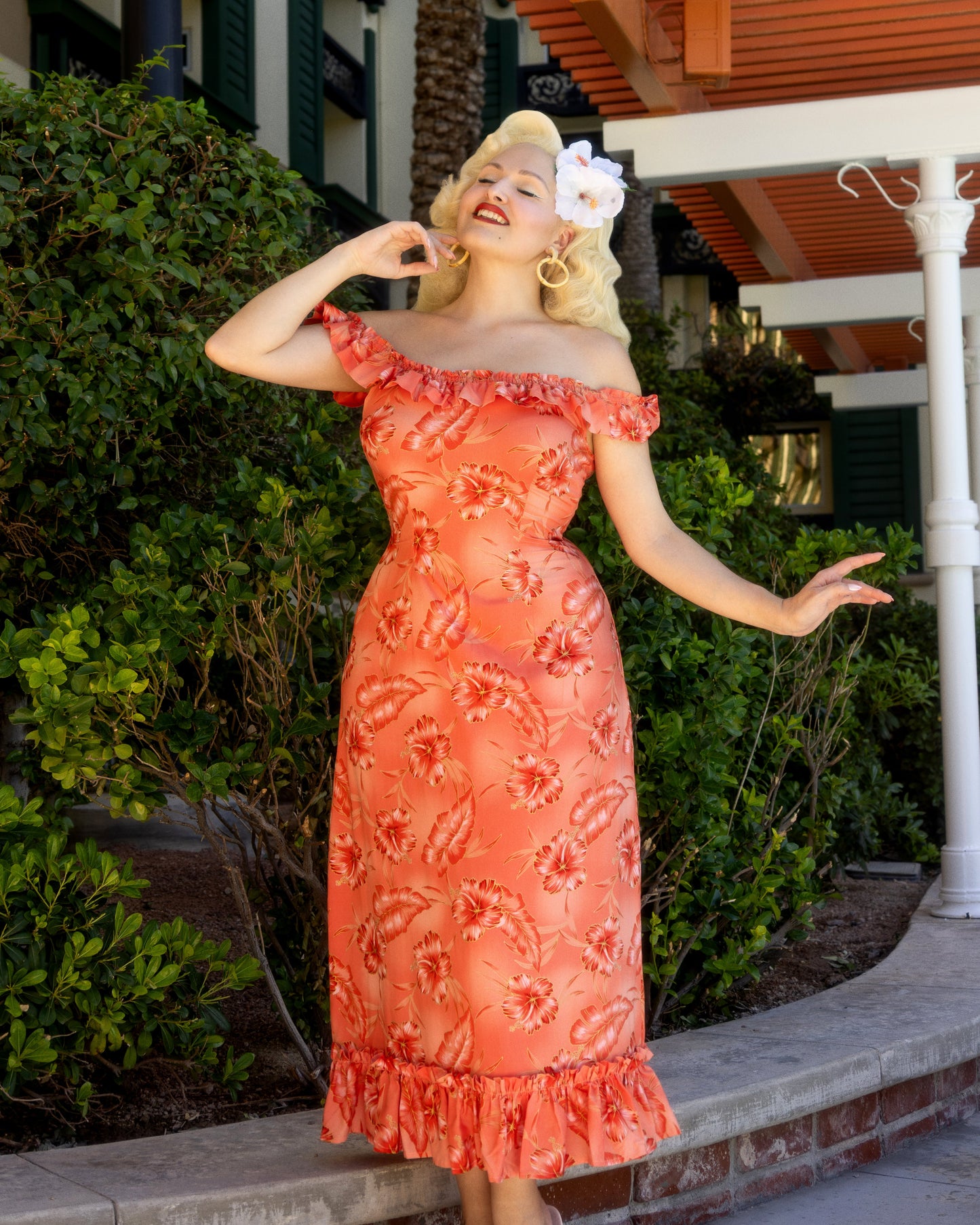 Woman in a coral lace dress posing outdoors with greenery and a building in the background