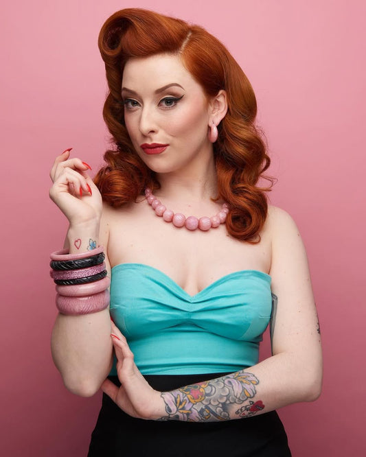 Woman with red hair and tattoos wearing a blue top against a pink background
