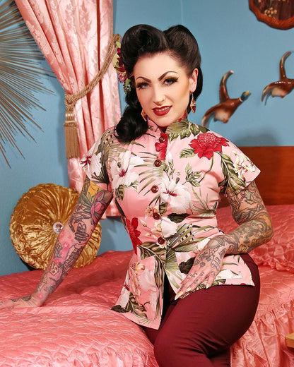 Woman with tattoos sitting on a bed in a room with pink and gold decor.